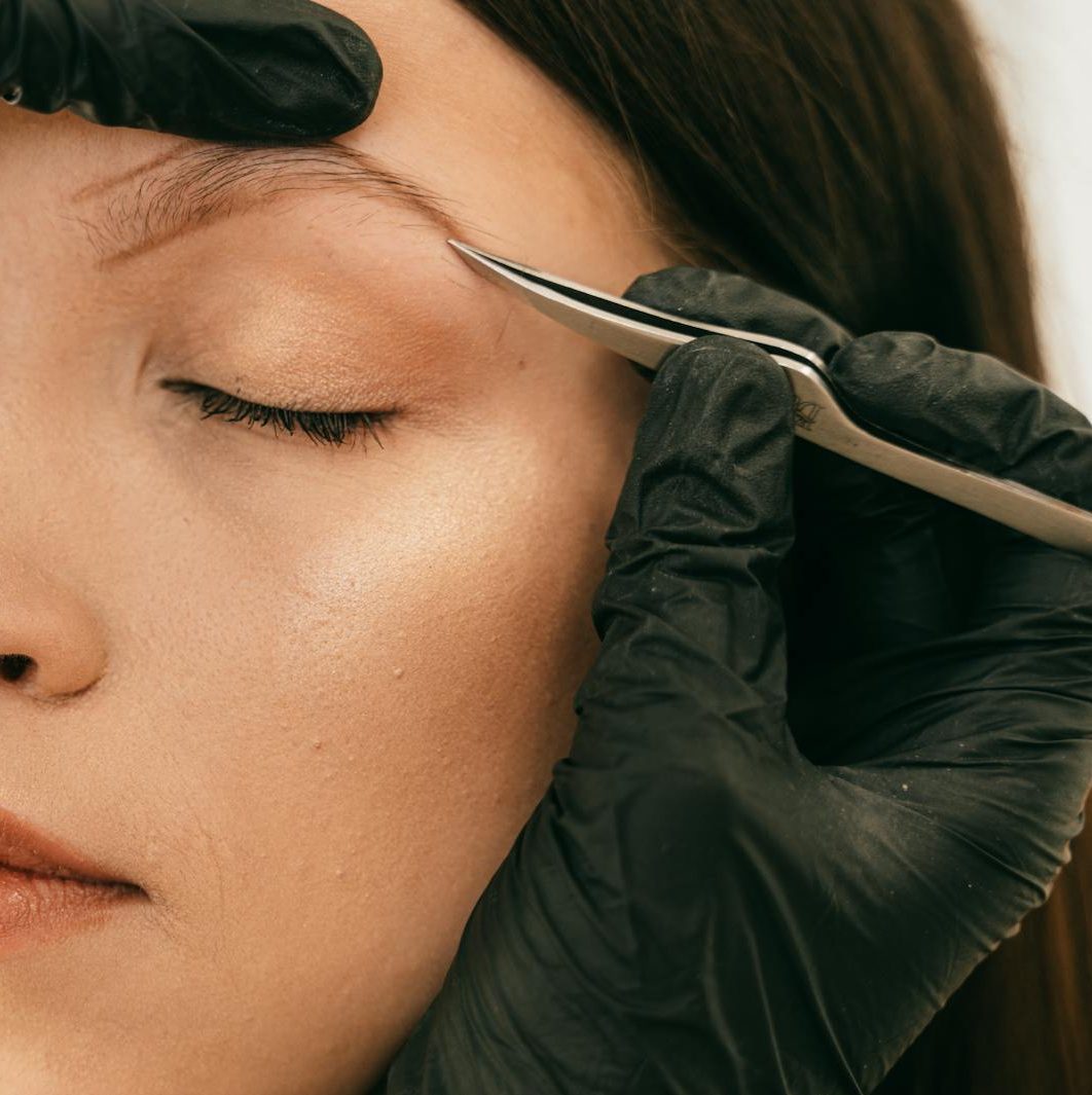 Detailed image of woman getting eyebrows plucked with tweezers by a beautician wearing gloves.